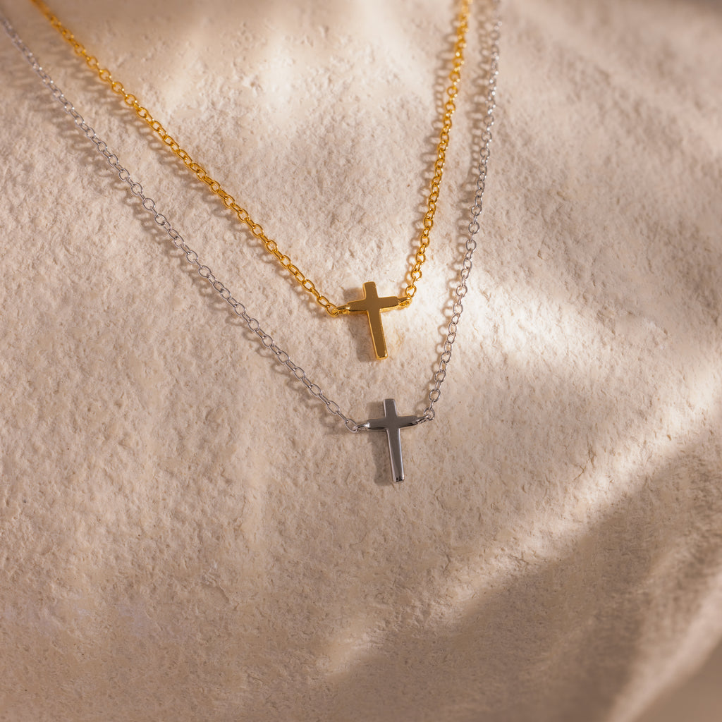 Two dainty cross charm necklaces in gold and silver displayed against a neutral stone surface.