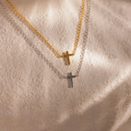 Two dainty cross charm necklaces in gold and silver displayed against a neutral stone surface.