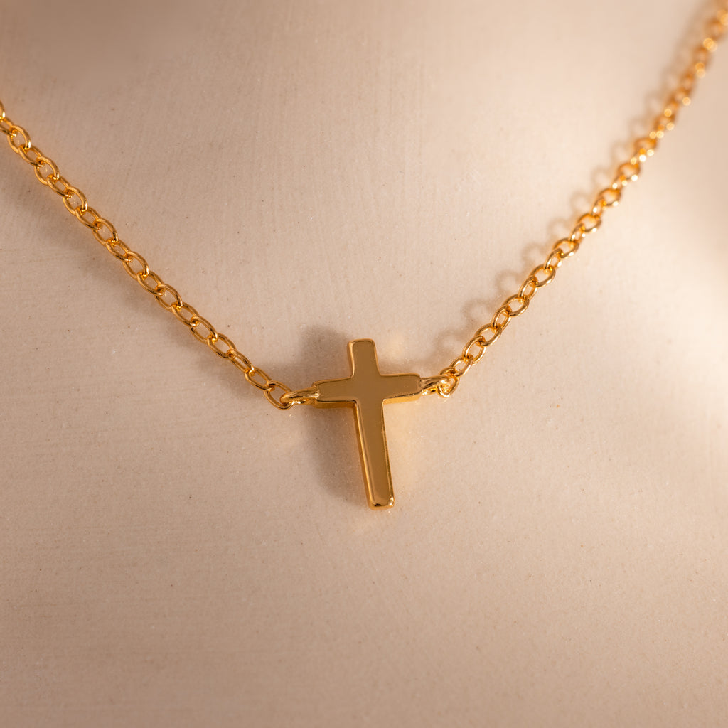 Close-up of a dainty gold cross charm necklace against a neutral surface.