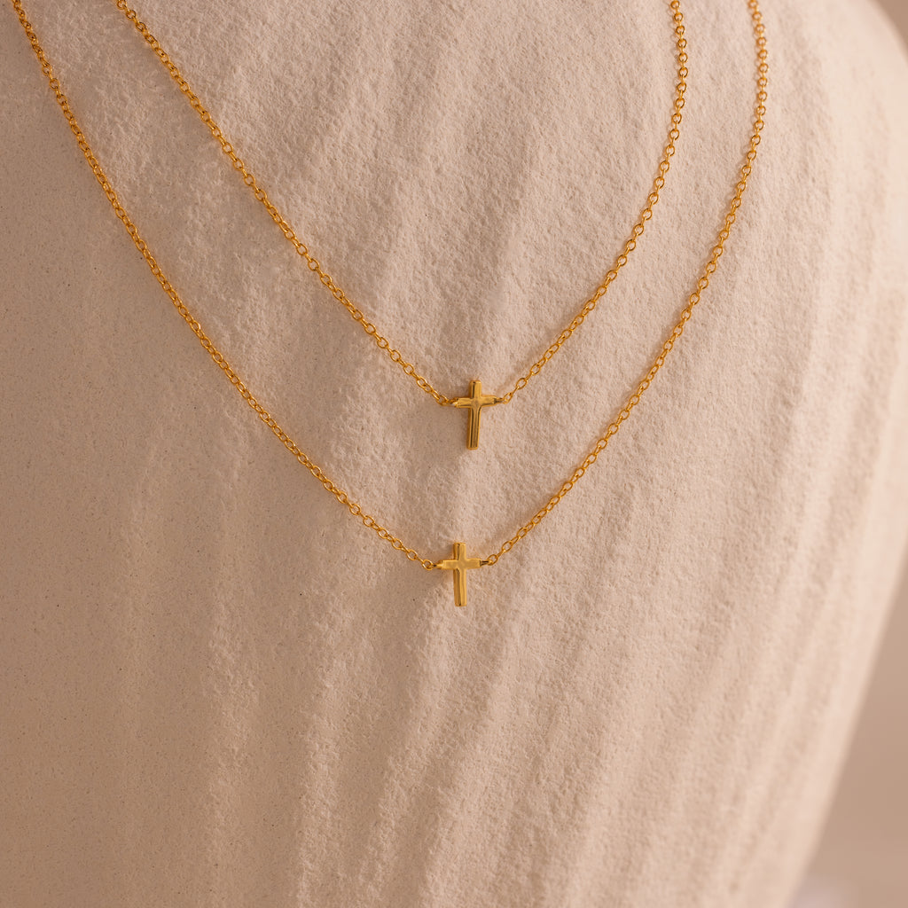 Two dainty gold cross charm necklaces in 16 and 18 inches, displayed against a neutral stone surface.