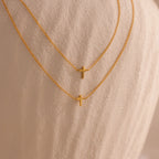 Two dainty gold cross charm necklaces in 16 and 18 inches, displayed against a neutral stone surface.