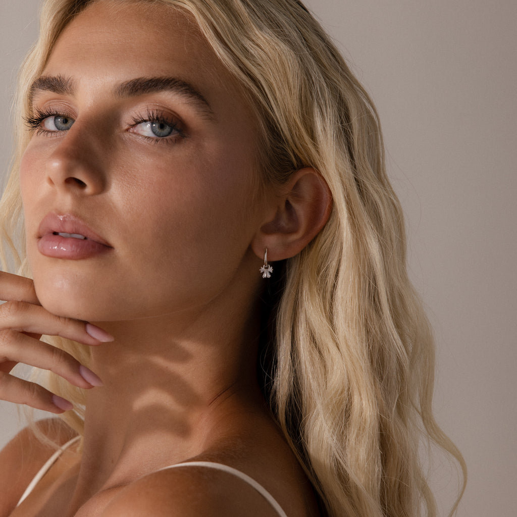 Close-up of silver hoop earrings with crystal-studded bow charms, displayed on a neutral background.
