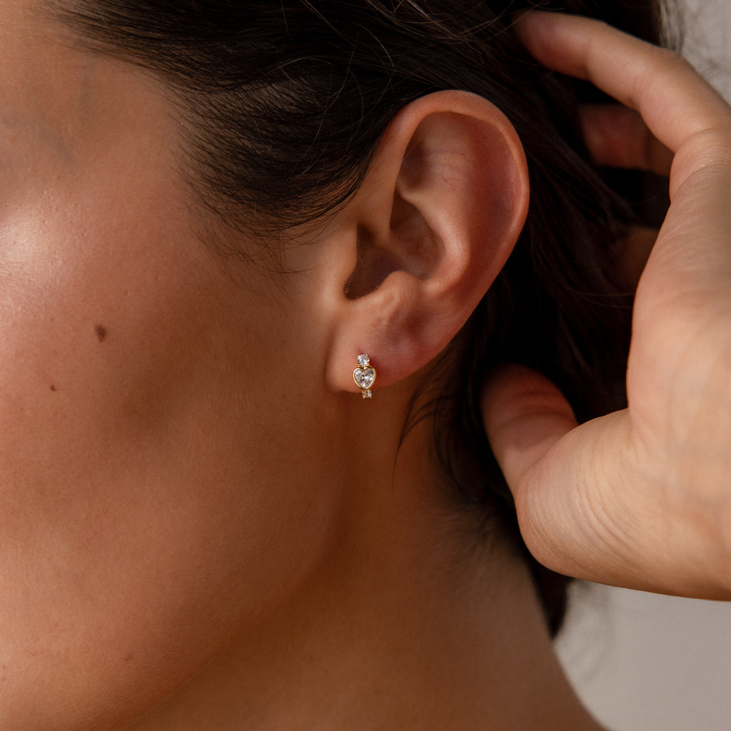 Close-up of a woman's ear wearing Birthstone Heart Hoops by Caitlyn Minimalist, featuring small, elegant heart-shaped earrings with red and clear gemstones.