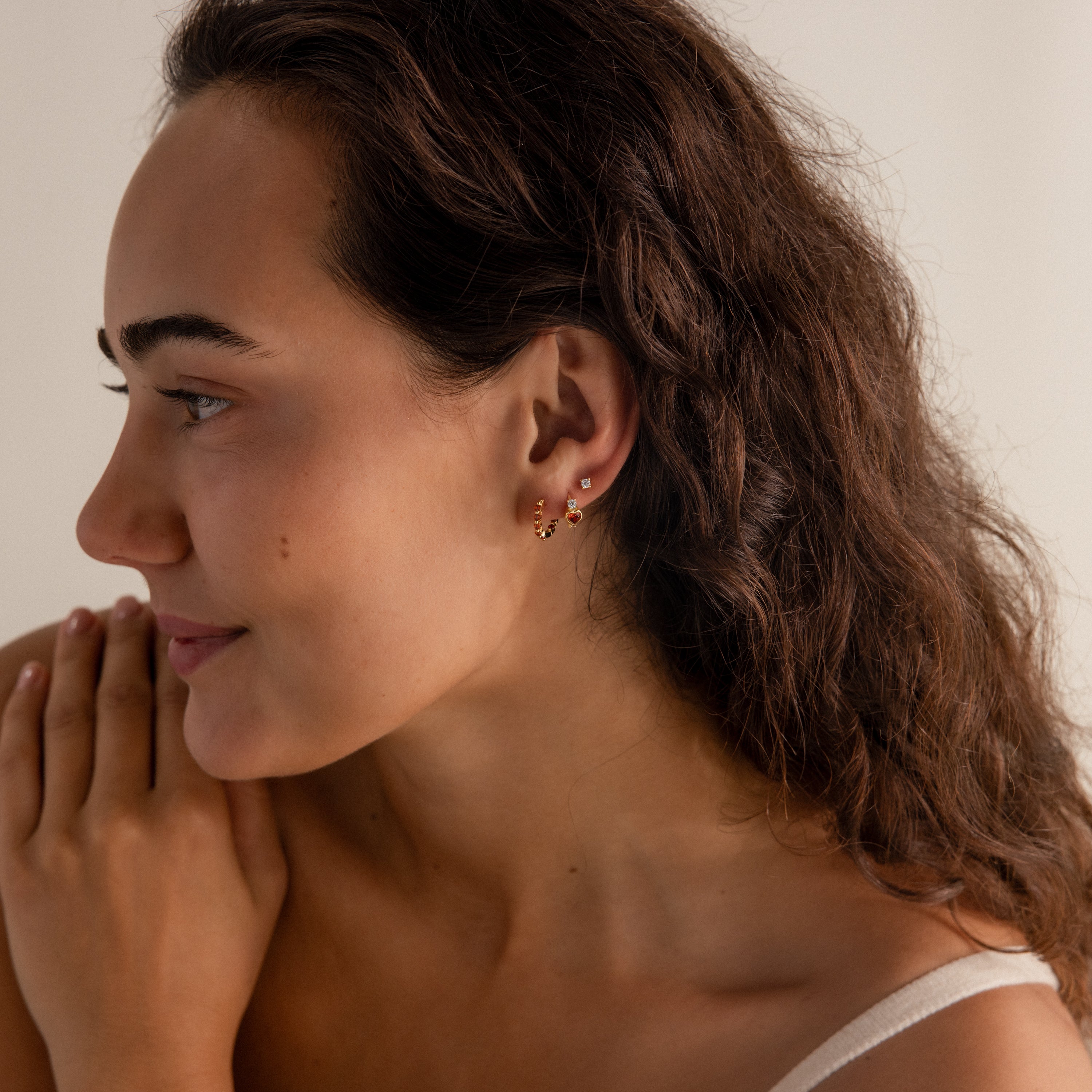 A woman with long brown hair, wearing Garnet Heart Hoops and a white top, looks to the left and smiles softly.