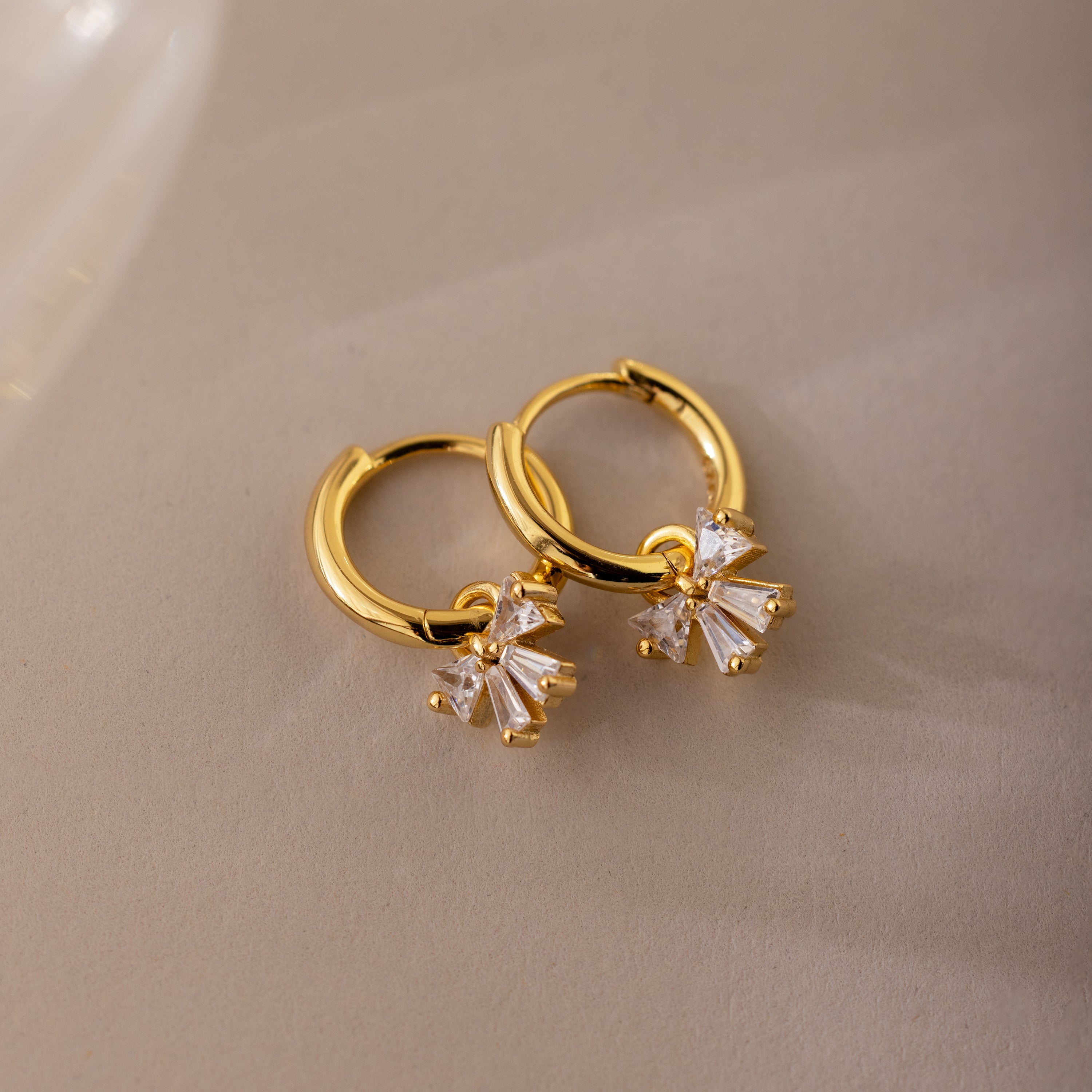 Gold Diamond Bow Hoop Earrings with delicate cubic zirconia bows, styled flat on a neutral taupe background.