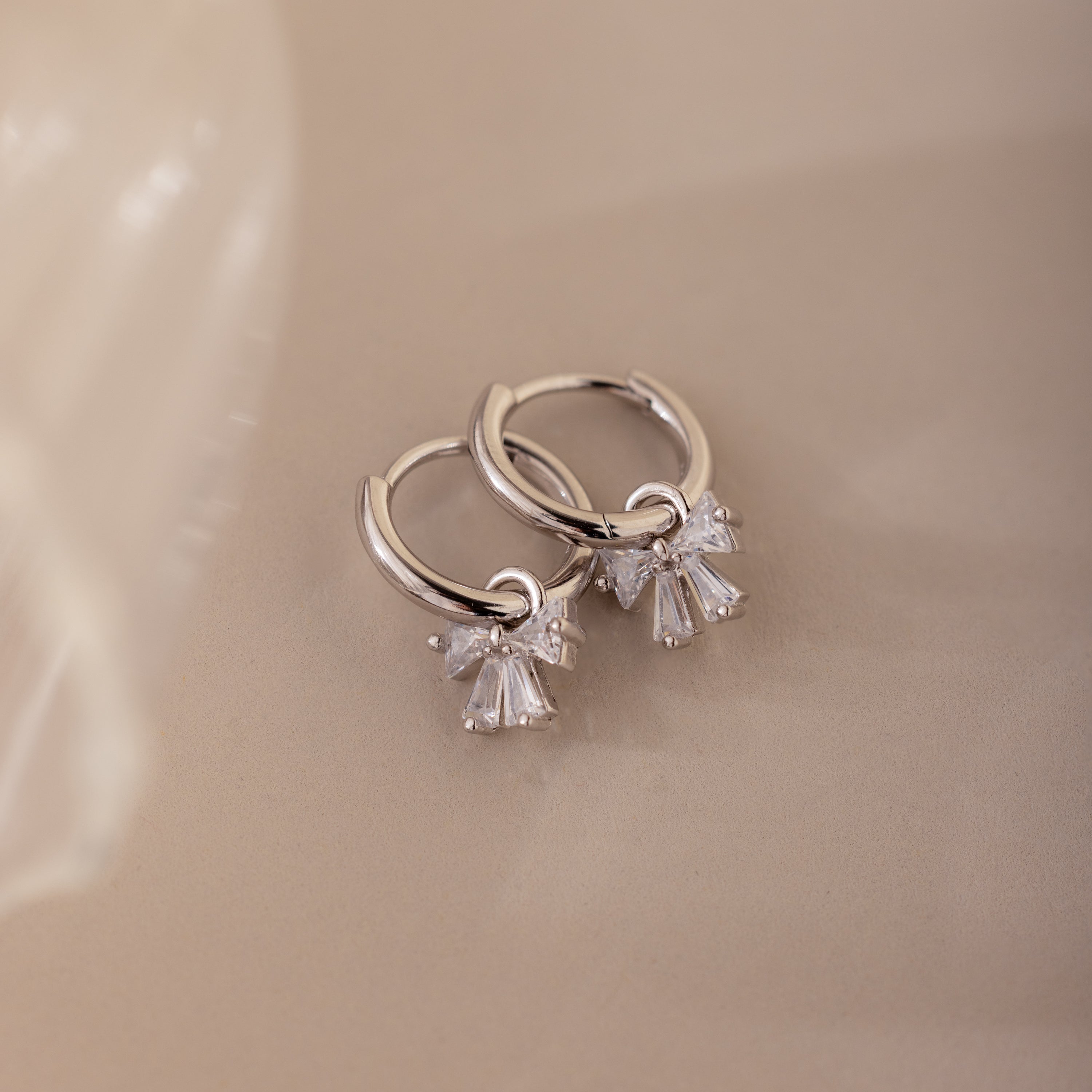 Close-up of silver hoop earrings with diamond-studded bow charms, displayed on a neutral background.