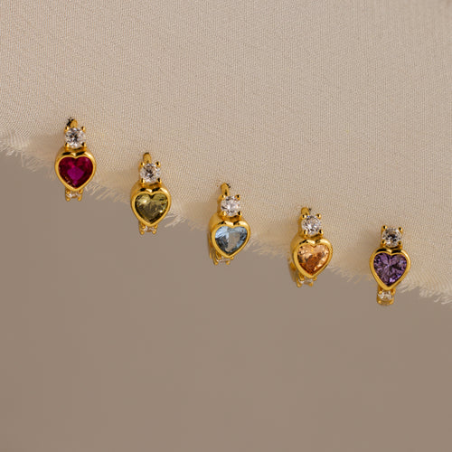 The Birthstone Heart Hoops by Caitlyn Minimalist feature heart-shaped gemstones in various colors, each accented with a clear stone—ideal for pairing with your favorite birthstone earrings or diamond huggies for a coordinated look.