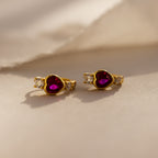A pair of Birthstone Heart Hoops by Caitlyn Minimalist, featuring heart-shaped Ruby gemstones and small clear stones, displayed on a soft beige surface.