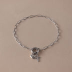 The Cross & Heart Charm Bracelet in Silver features a sleek paperclip chain with delicate cross and heart charms, shown on a plain beige background.