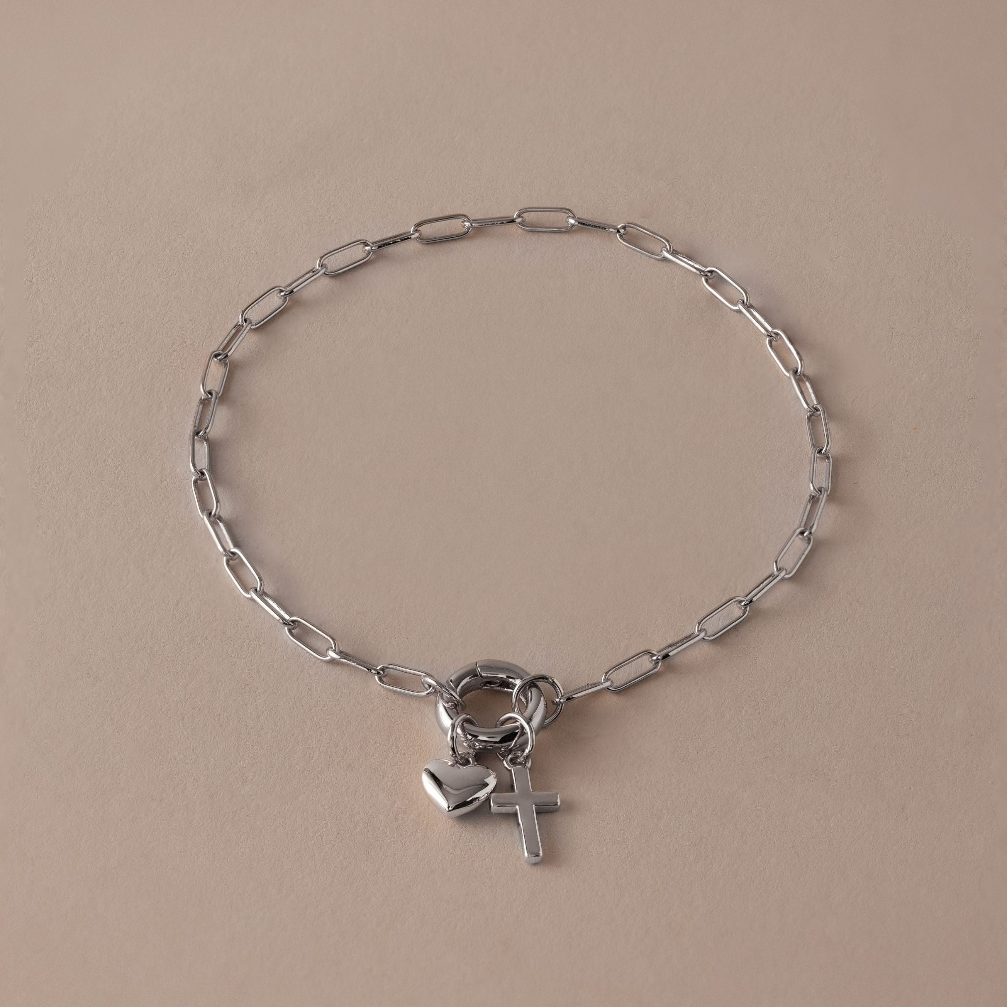 The Cross & Heart Charm Bracelet in Silver features a sleek paperclip chain with delicate cross and heart charms, shown on a plain beige background.