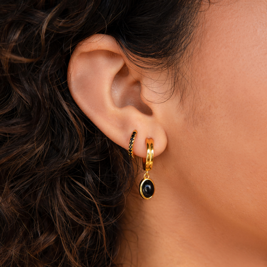 Close-up of a woman's ear adorned with three gold earrings from the Black Onyx Hoops Bundle, featuring a striking black stone, with her curly hair softly framing the look.