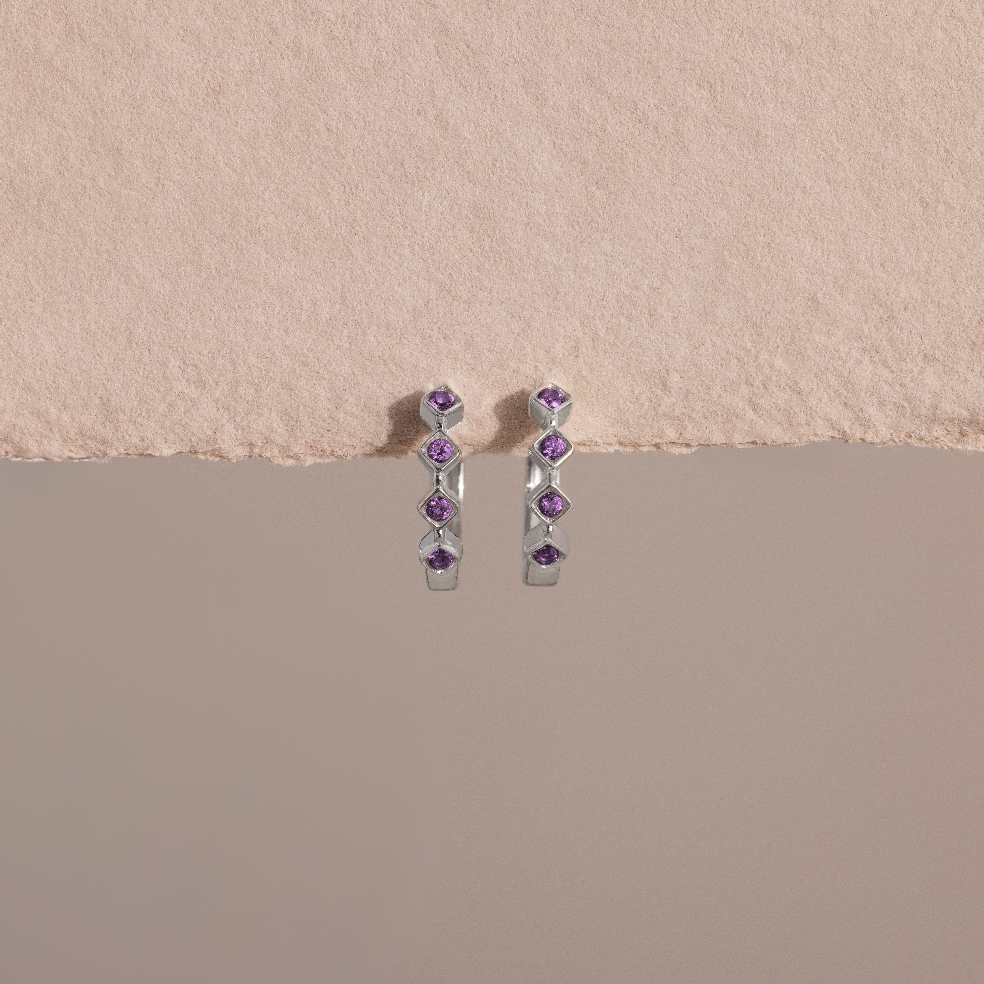 Geometric Amethyst Huggies in silver, each adorned with five small amethyst stones and subtle diamond accents, displayed against a textured beige background.