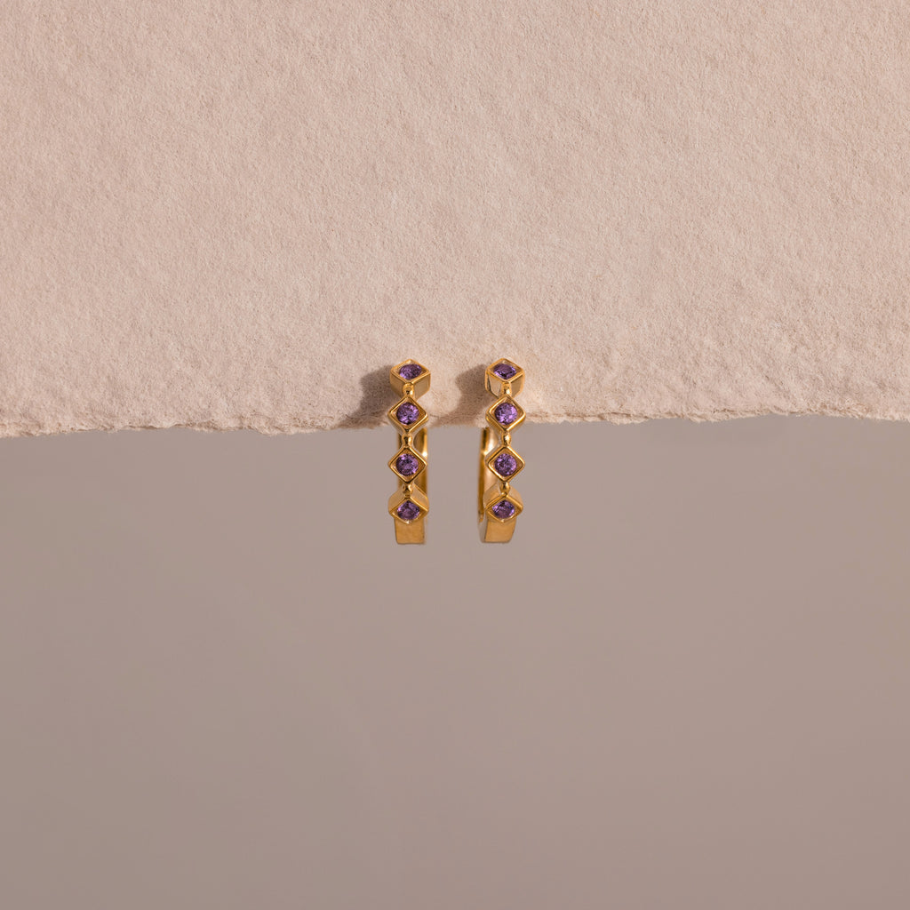 Geometric Amethyst Huggies—gold earrings with vertically set amethyst stones and subtle diamond accents—displayed on a beige textured background.