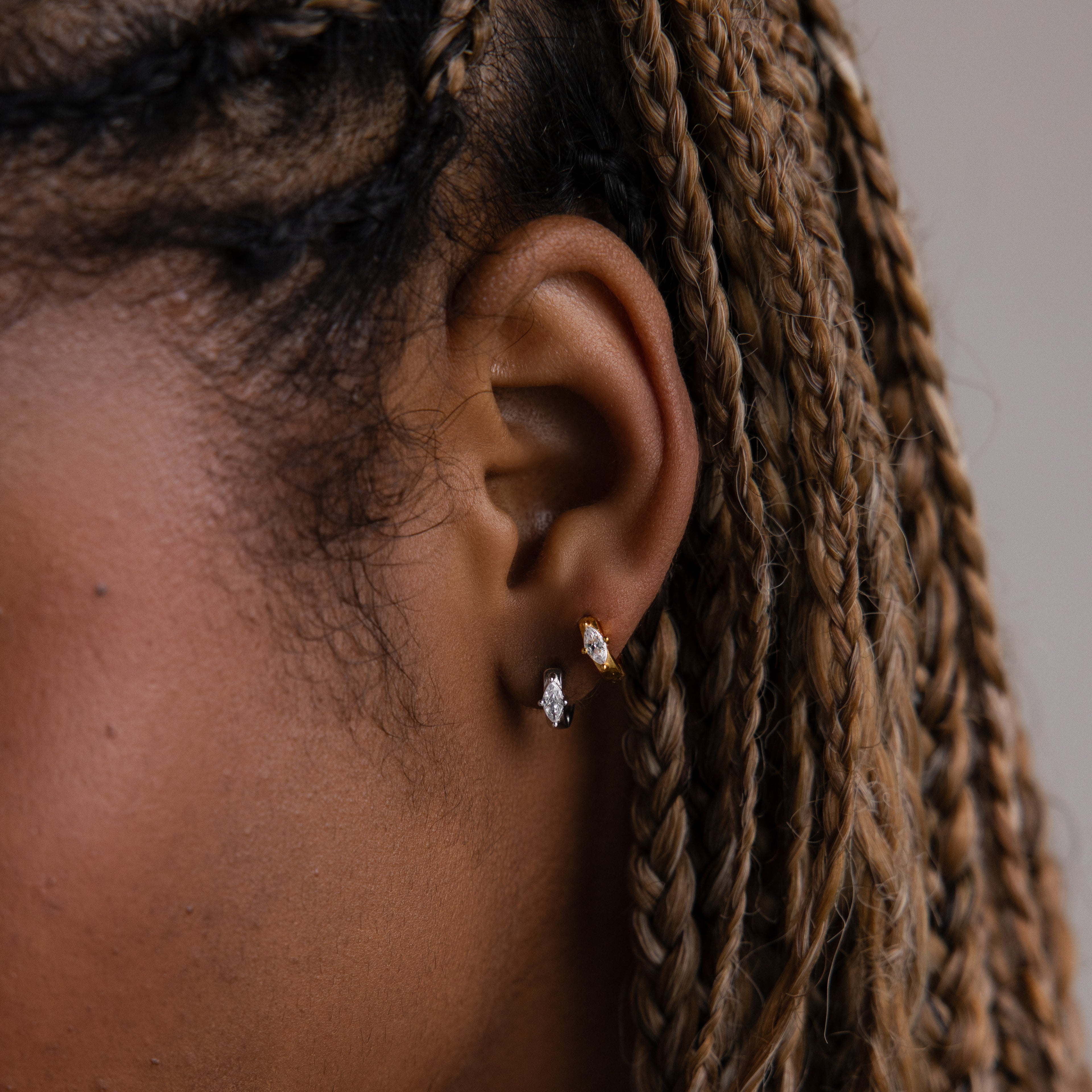 Close-up of a model with braided hair and gold and silver diamond huggie hoop earrings against a neutral background