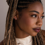 Close-up of a woman with braided hair wearing silver diamond stud earrings and a white top against a neutral background