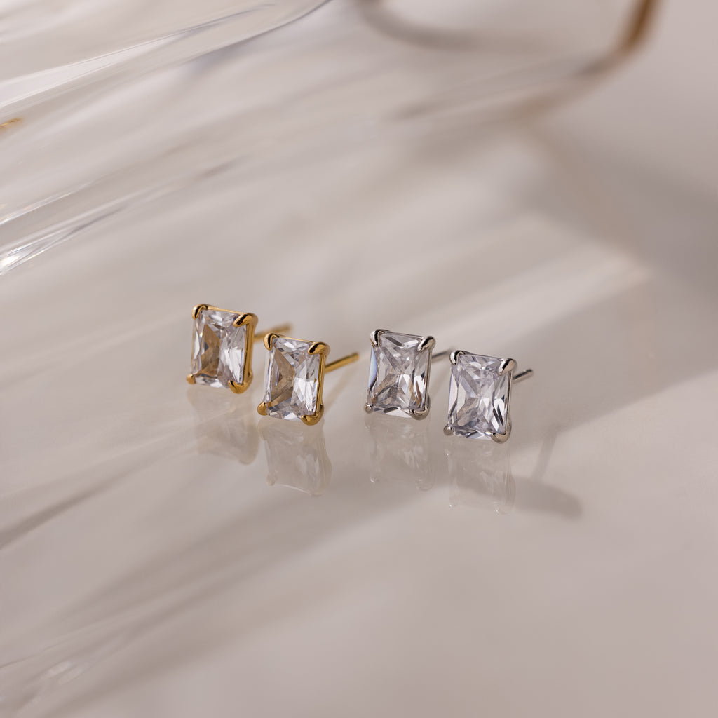 Two pairs of gold and silver stud earrings with clear gemstones on a light background