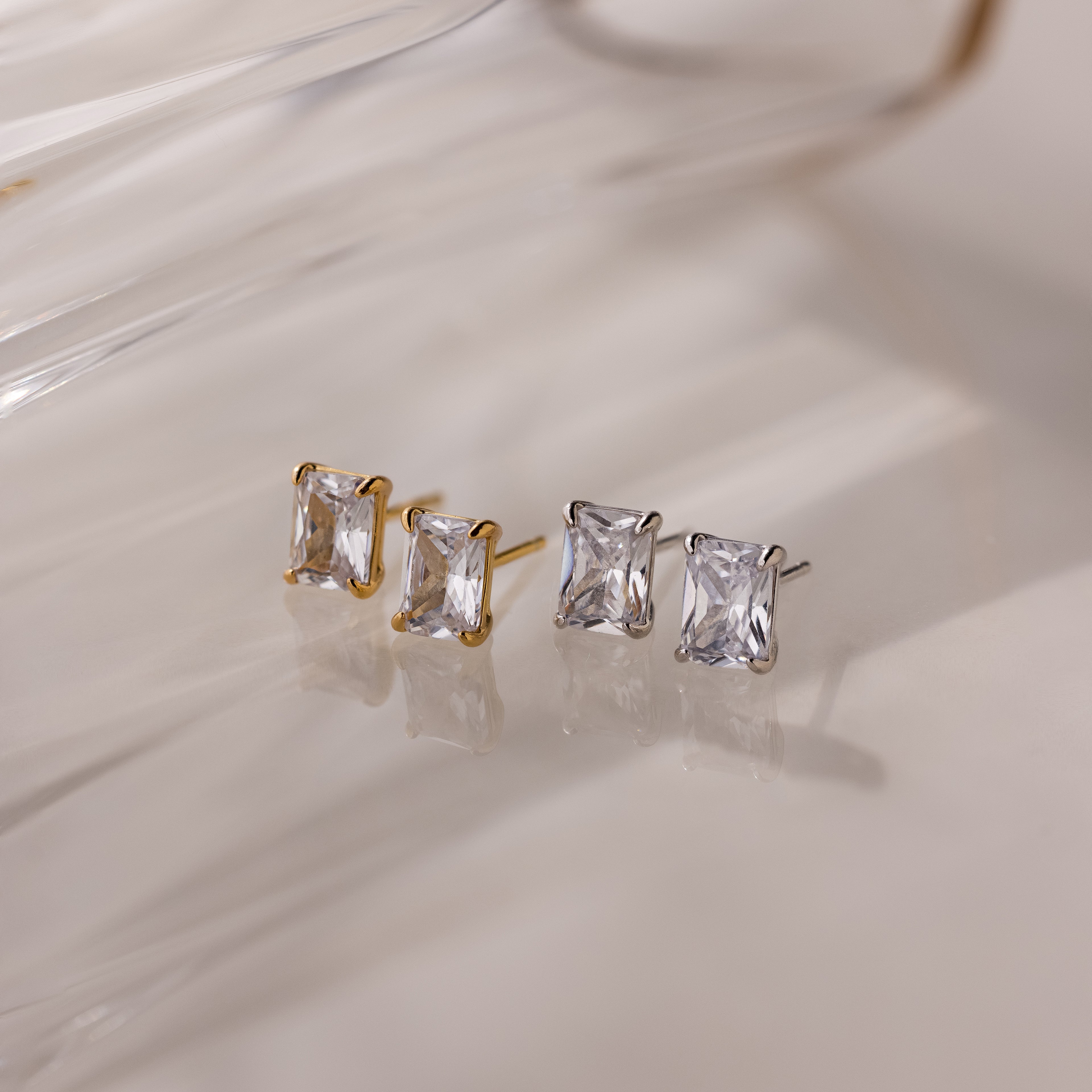 Two pairs of gold and silver stud earrings with clear gemstones on a light background