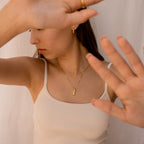 Woman in a cream tank top wears layered gold necklaces, including the standout Kinn Body Necklace, as she raises her hands to partially cover her face.