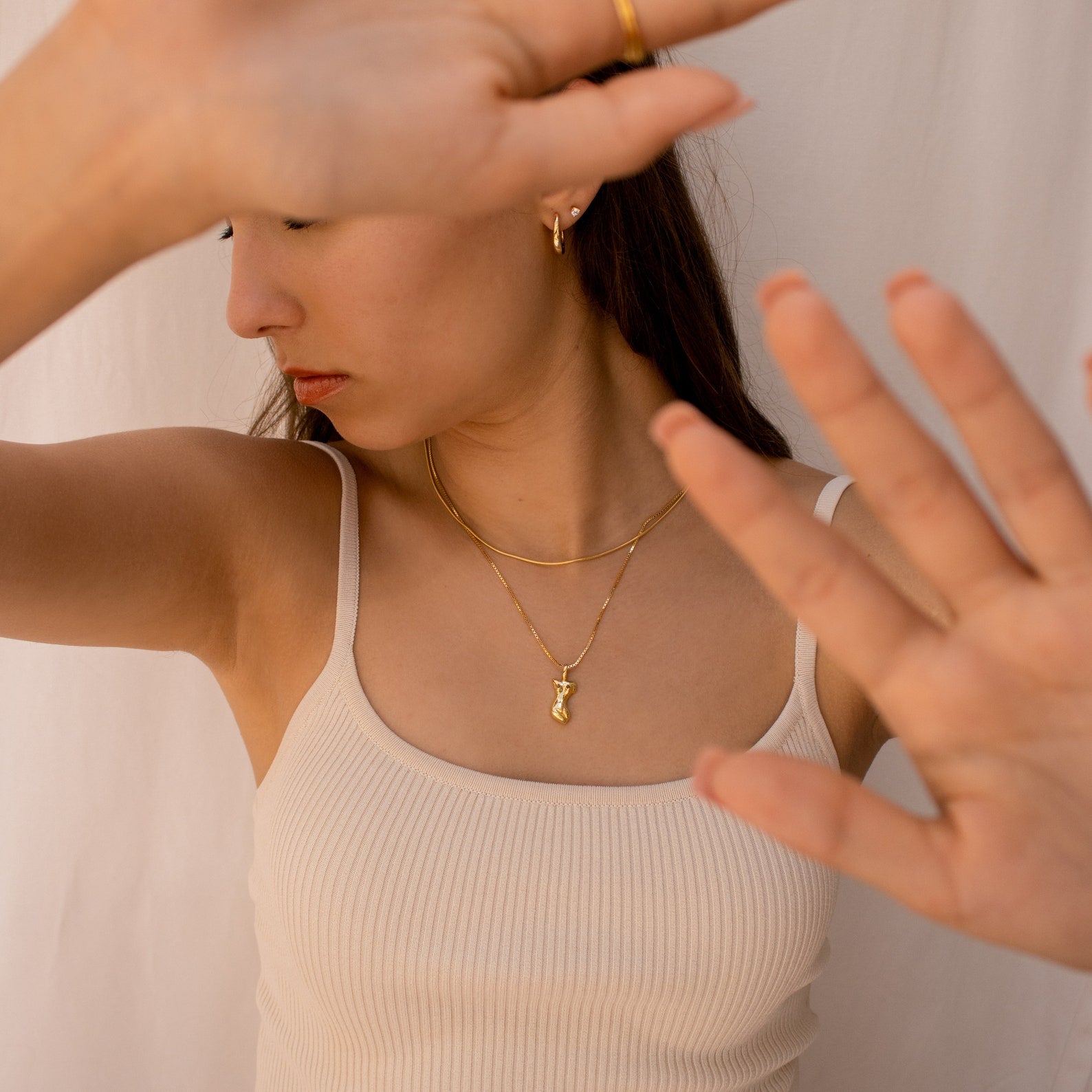 Woman in a cream tank top wears layered gold necklaces, including the standout Kinn Body Necklace, as she raises her hands to partially cover her face.