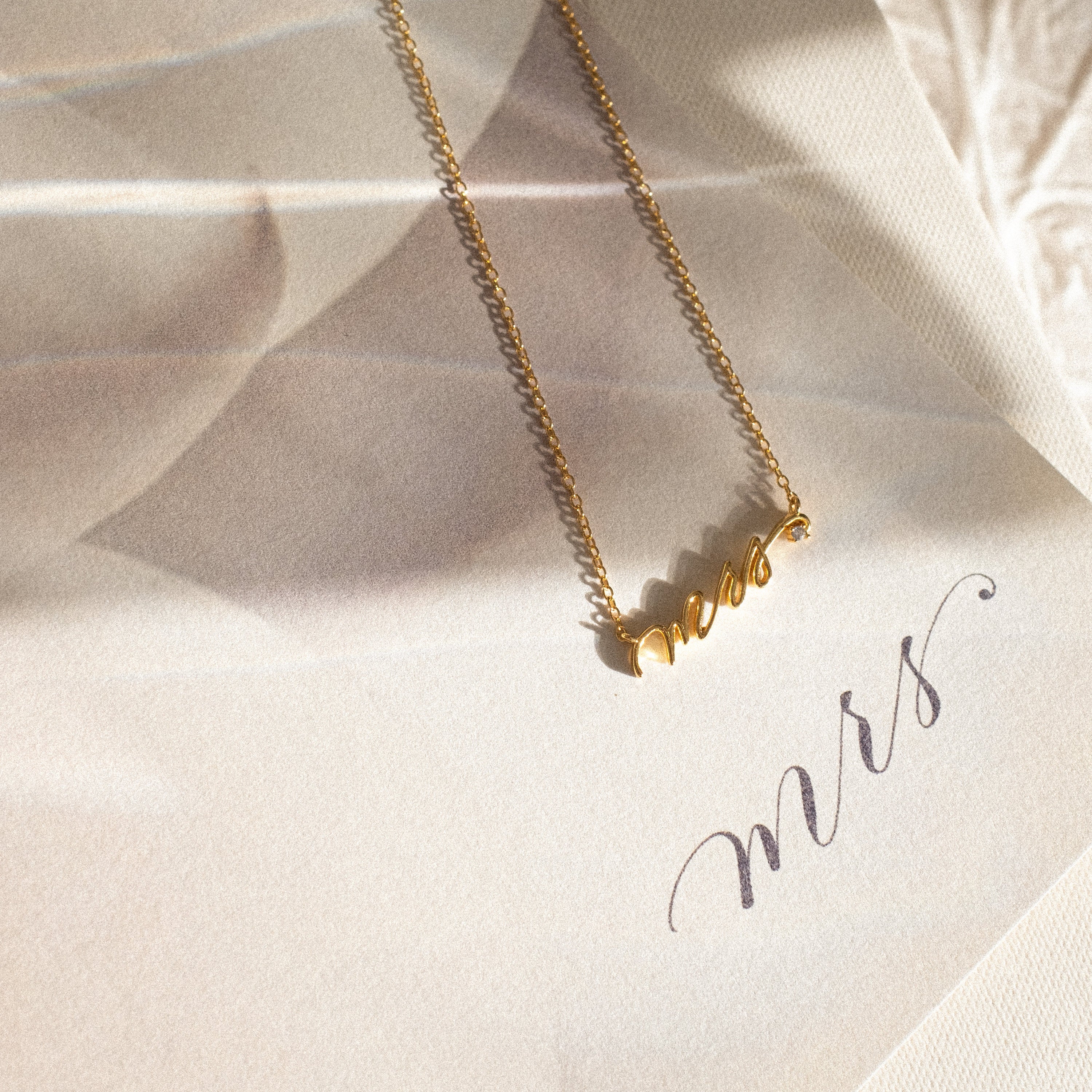 The Mrs. Necklace glows in soft, natural sunlight with “mrs” written in cursive—an elegant wedding gift for the bride-to-be.
