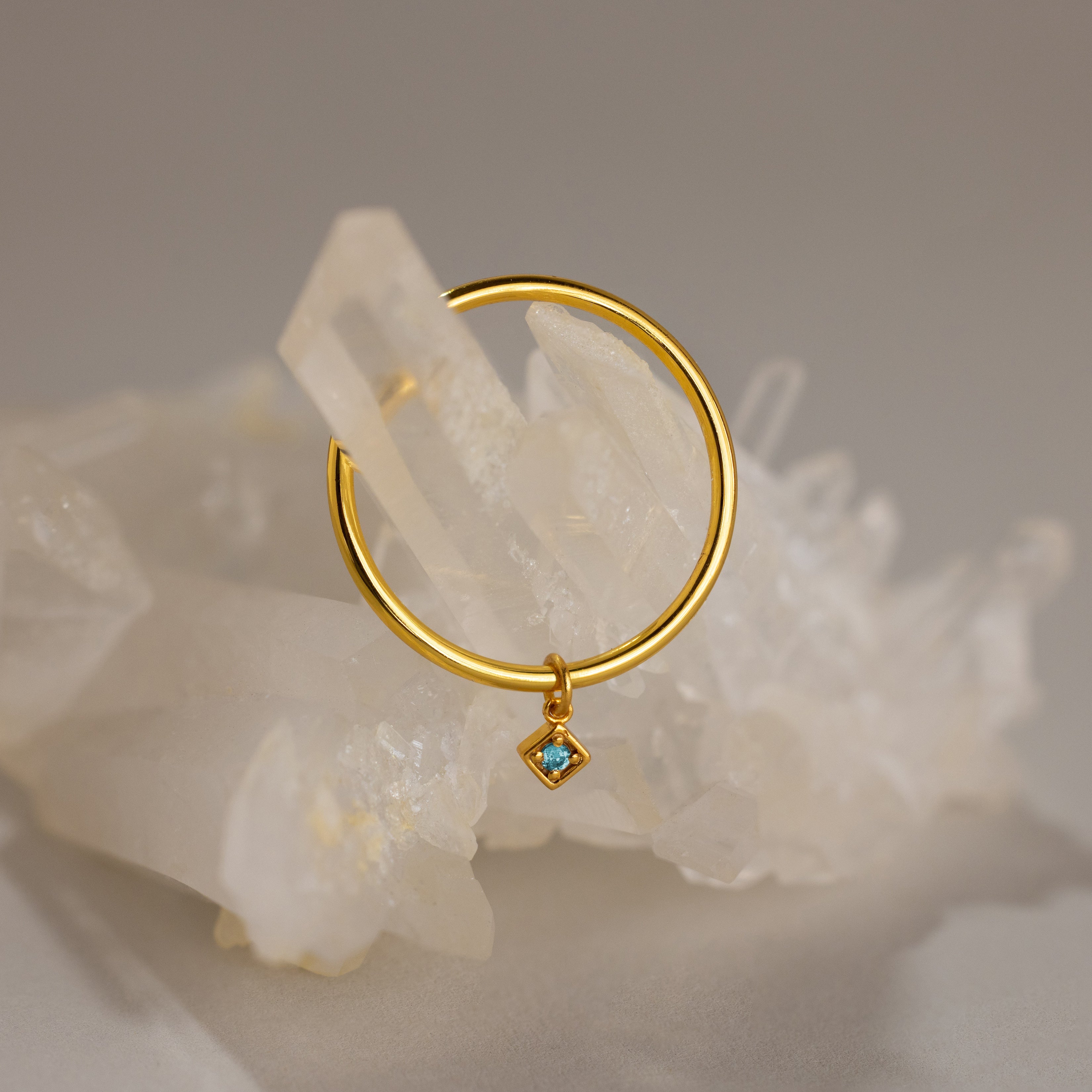 The Dainty Birthstone Drop Ring features a gold hoop with a small blue gemstone charm, shown on a clear quartz crystal cluster—an ideal personalized jewelry piece for birthstone lovers.