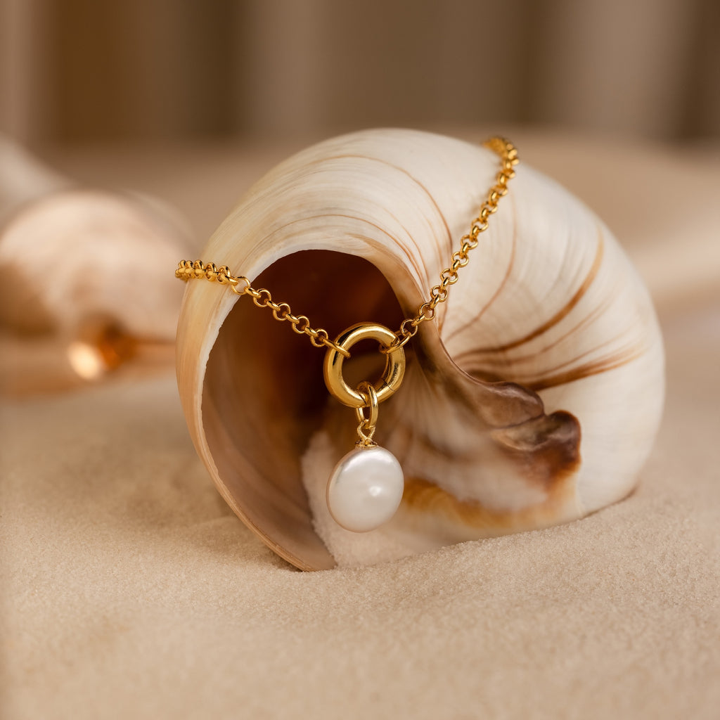 The Round Pearl Charm in 18K Gold, featuring a round pearl and Mother of Pearl pendant, is elegantly draped over a seashell resting on sand.