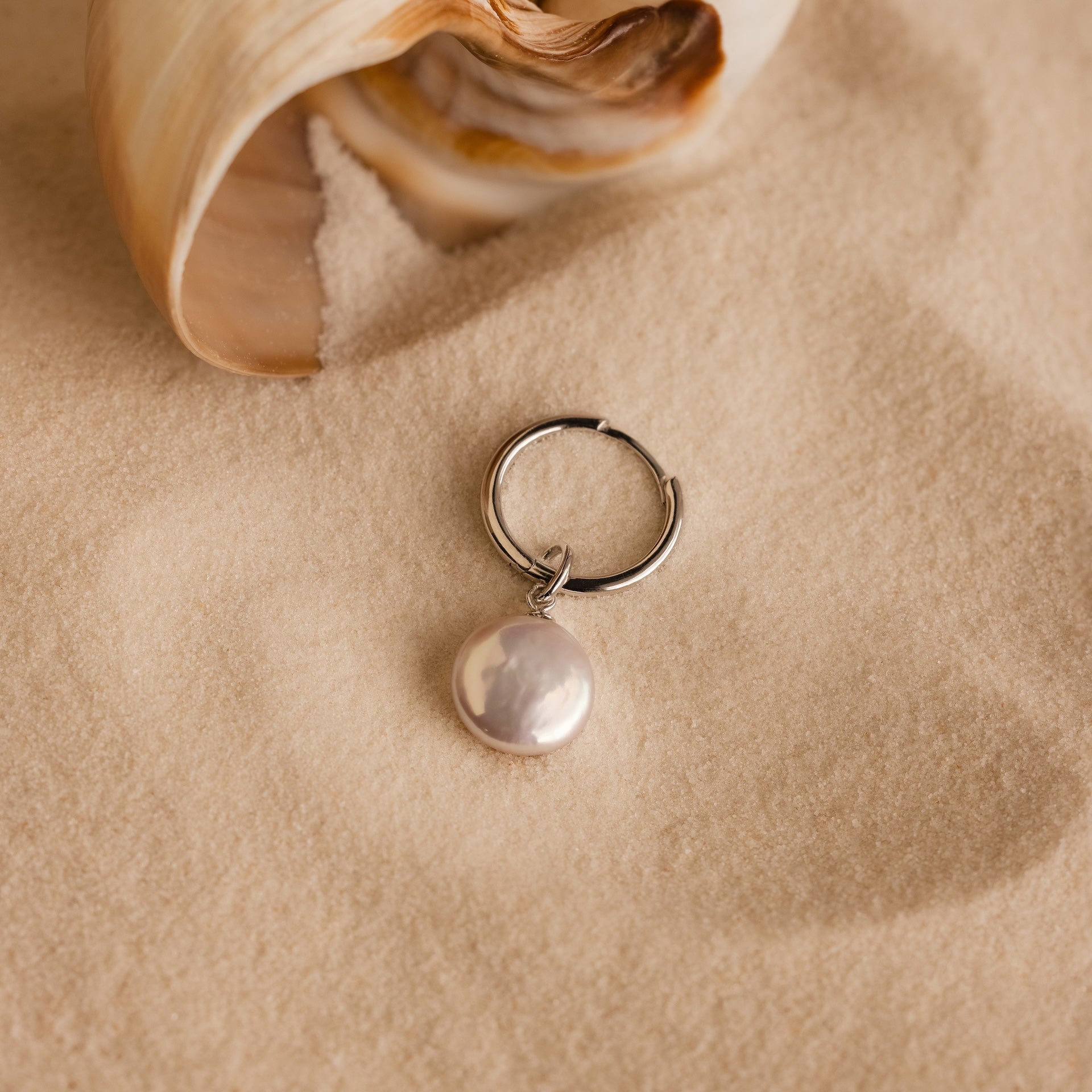 The Round Pearl Charm in Silver rests on sand near a seashell, highlighting elegant coastal-inspired jewelry.