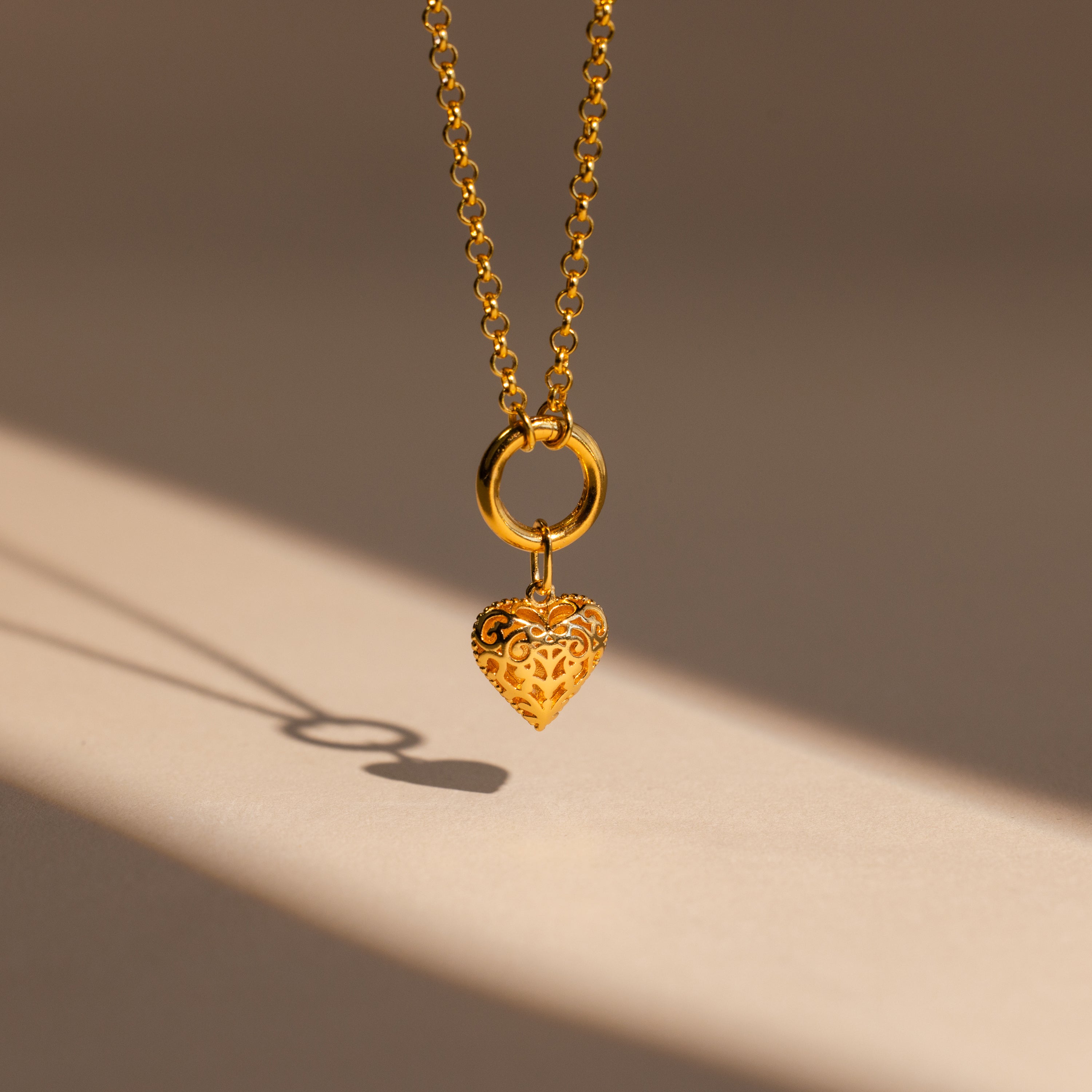 The Vintage Heart Charm necklace features a delicate gold chain and ornate heart pendant, casting a shadow on beige—an elegant addition to your jewelry collection or the perfect anniversary gift.