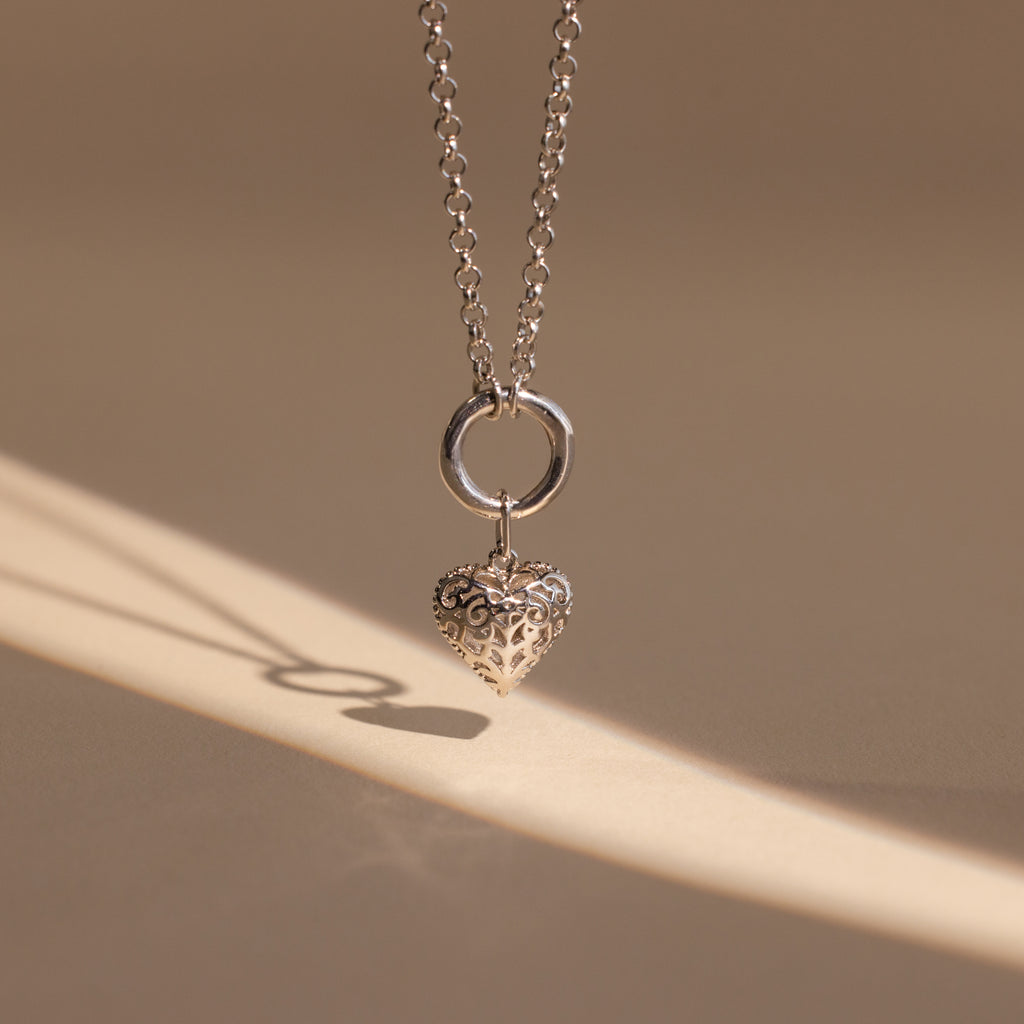 The Vintage Heart Charm in Sterling Silver casts a shadow on a beige surface—a timeless necklace, ideal for your jewelry collection or as an anniversary gift.