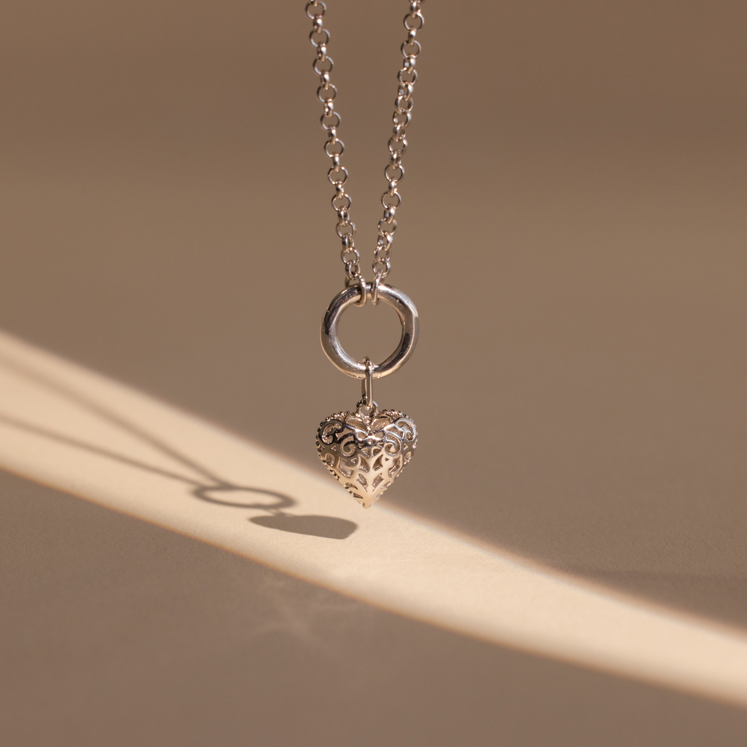 The Vintage Heart Charm in Sterling Silver casts a shadow on a beige surface—a timeless necklace, ideal for your jewelry collection or as an anniversary gift.