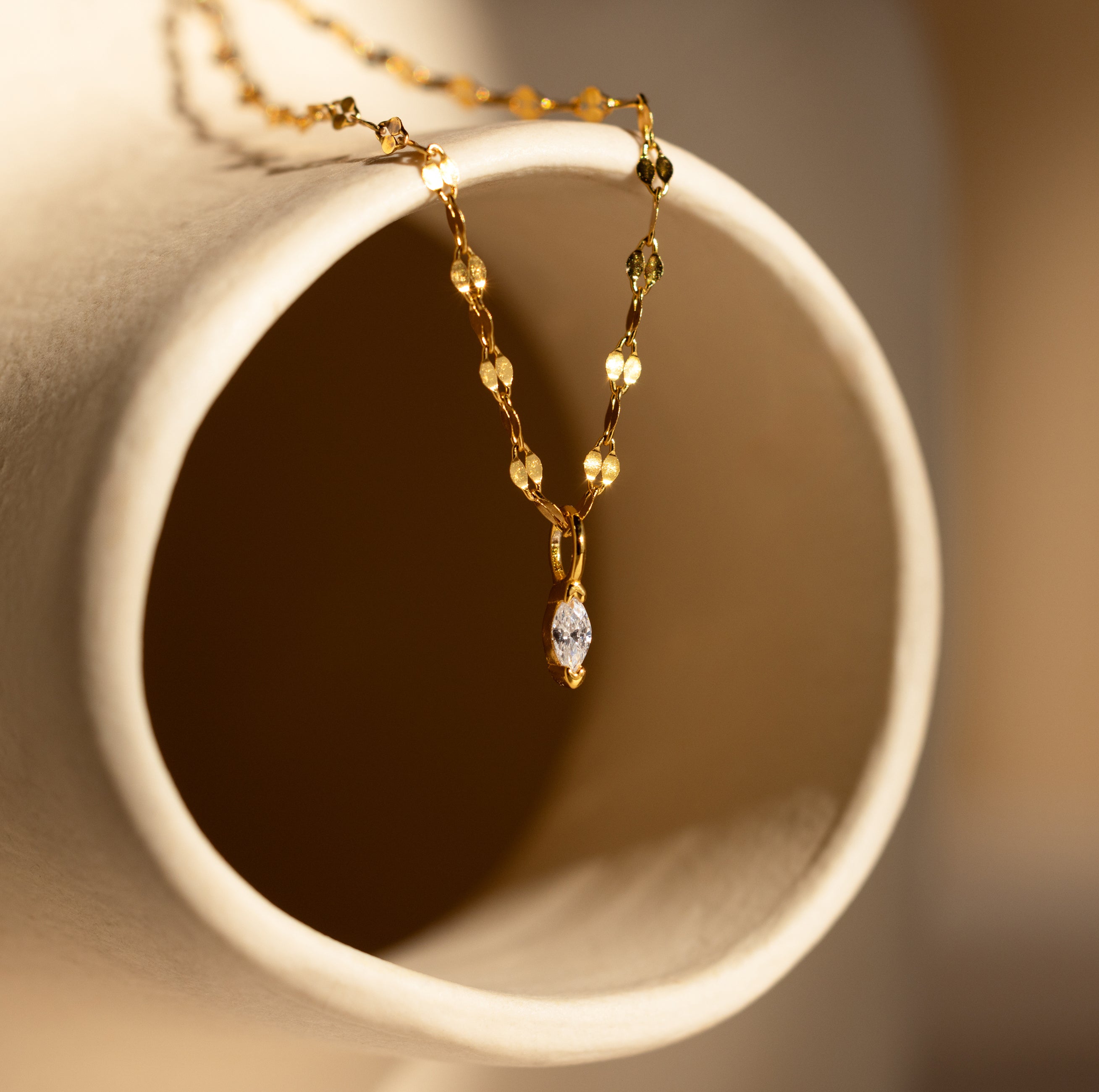 The Dainty Marquise Pendant Necklace drapes elegantly over a curved beige ceramic surface, highlighting its marquise-cut diamond charm on a delicate mirror twist chain.