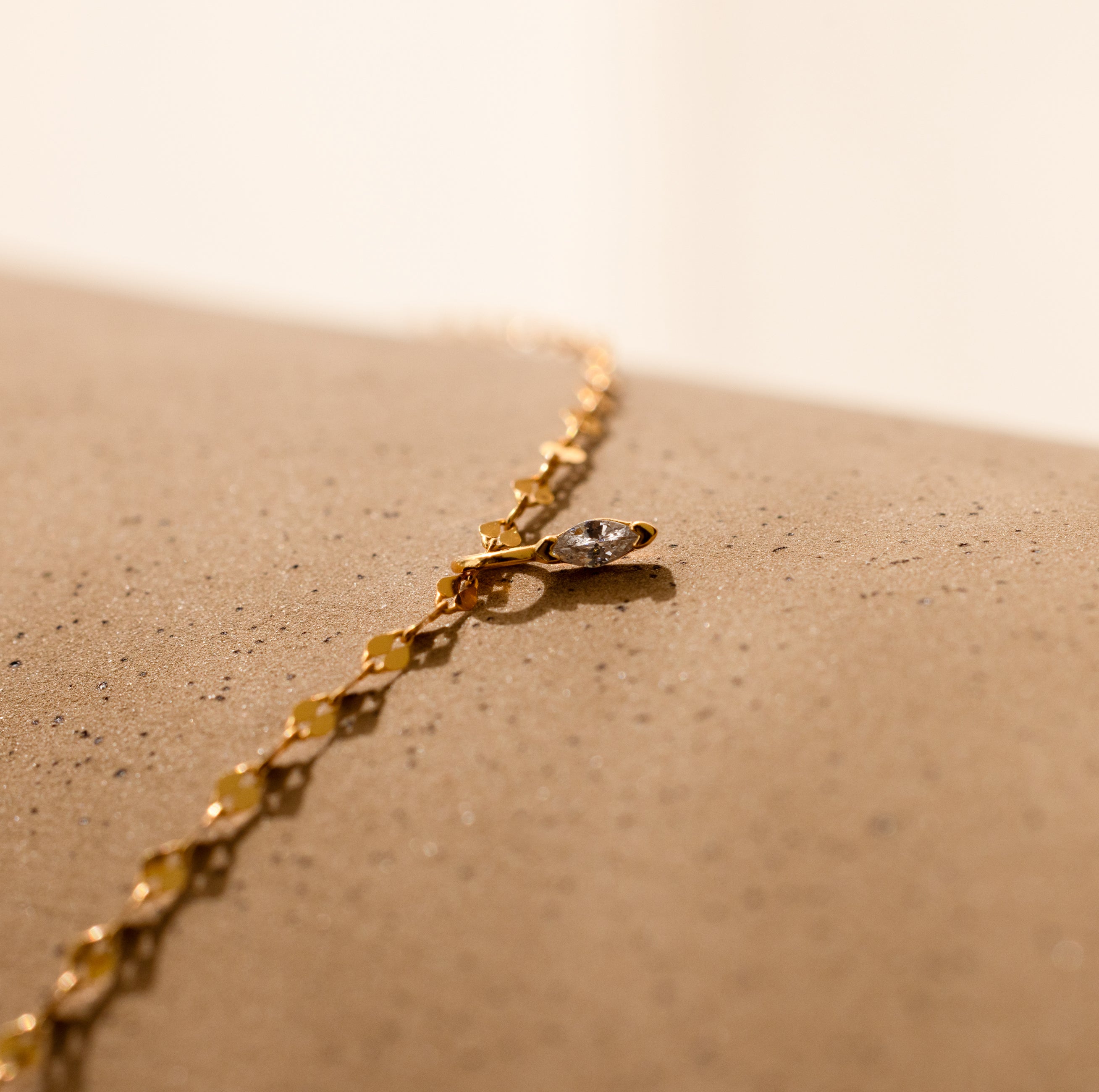A Dainty Marquise Pendant Necklace in 18K Gold is displayed on a textured beige surface.