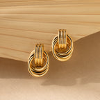 Triple Knot Earrings—gold hoops with interlocking rings—are shown on a beige textured background and folded fabric, offering an elegant earring choice and a perfect anniversary gift.