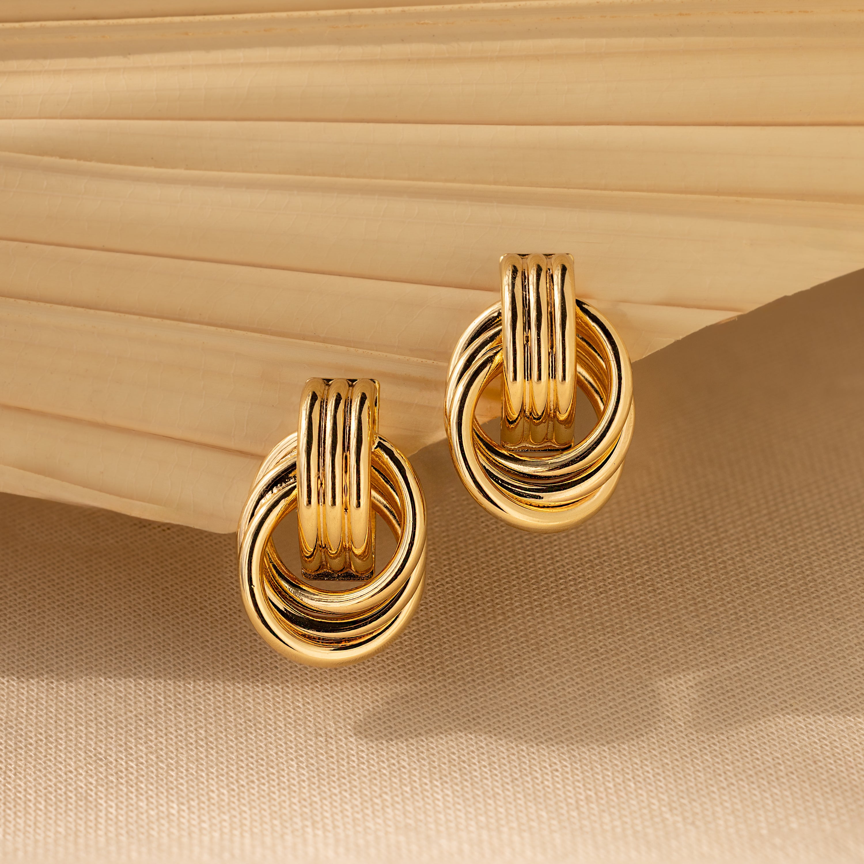 Triple Knot Earrings—gold hoops with interlocking rings—are shown on a beige textured background and folded fabric, offering an elegant earring choice and a perfect anniversary gift.