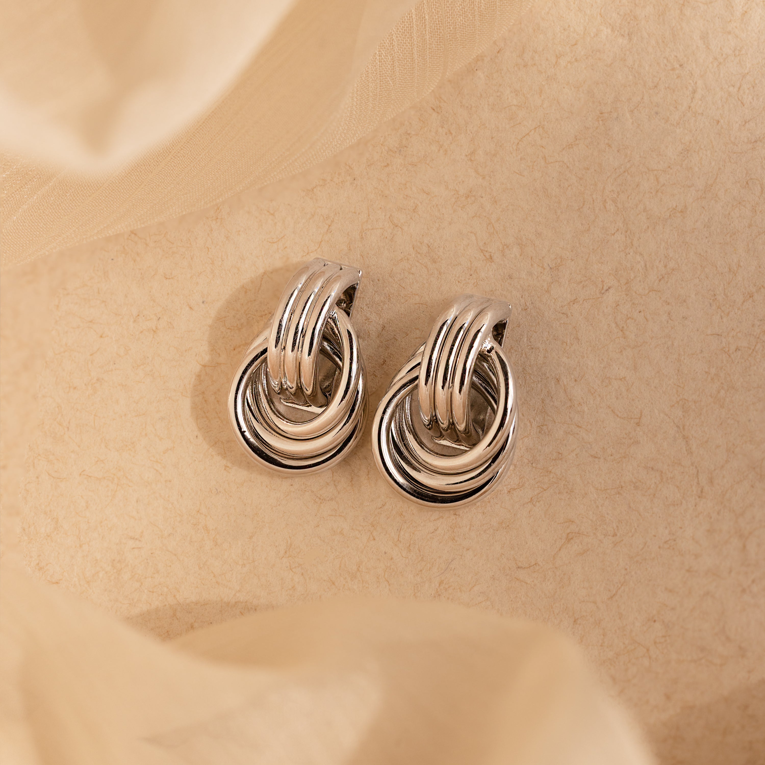 The Triple Knot Earrings in Silver feature interlocking loops and are beautifully displayed on a textured beige background with soft fabric accents—an ideal anniversary gift.