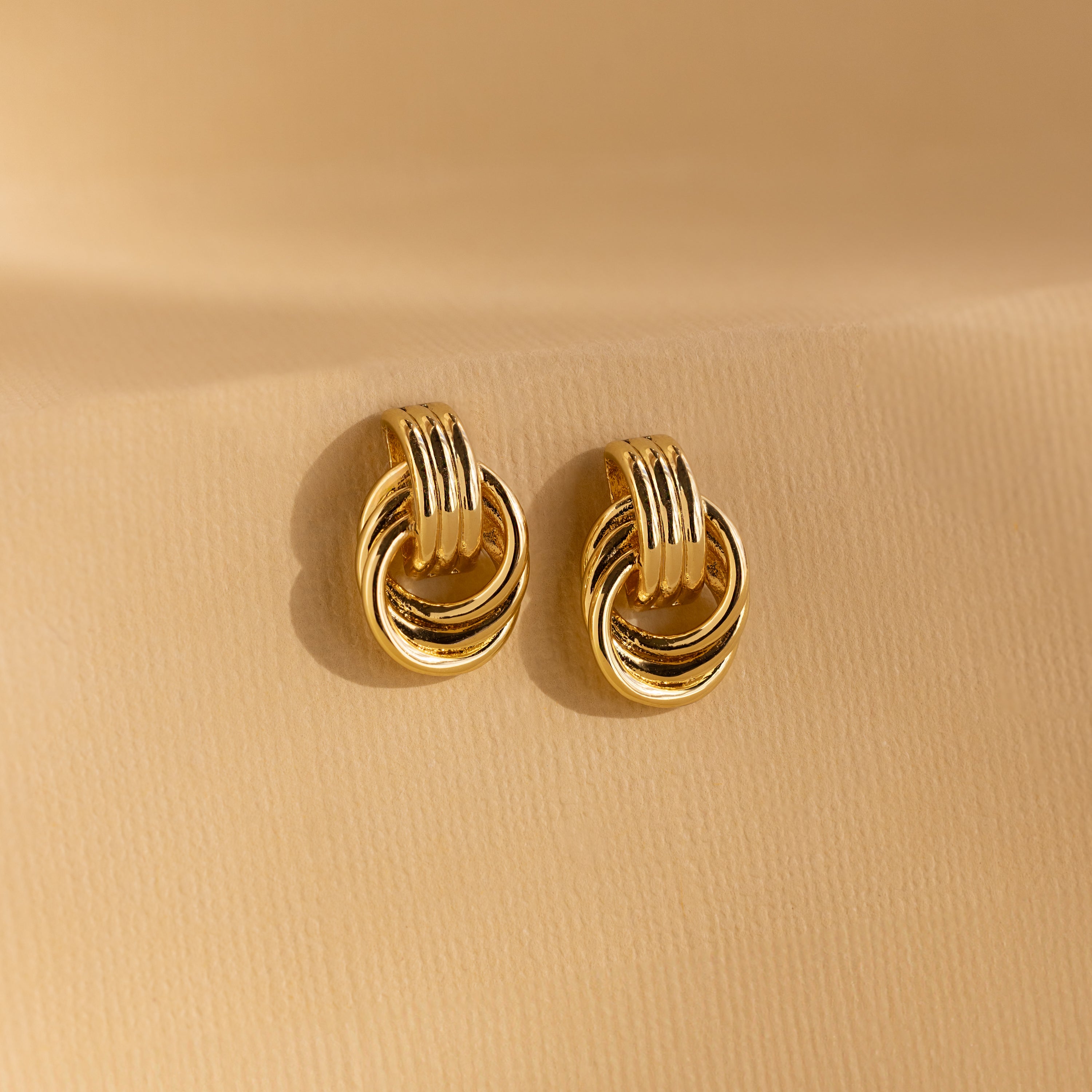 A pair of Mini Triple Knot Earrings in gold displayed upright on a smooth, neutral-toned surface.