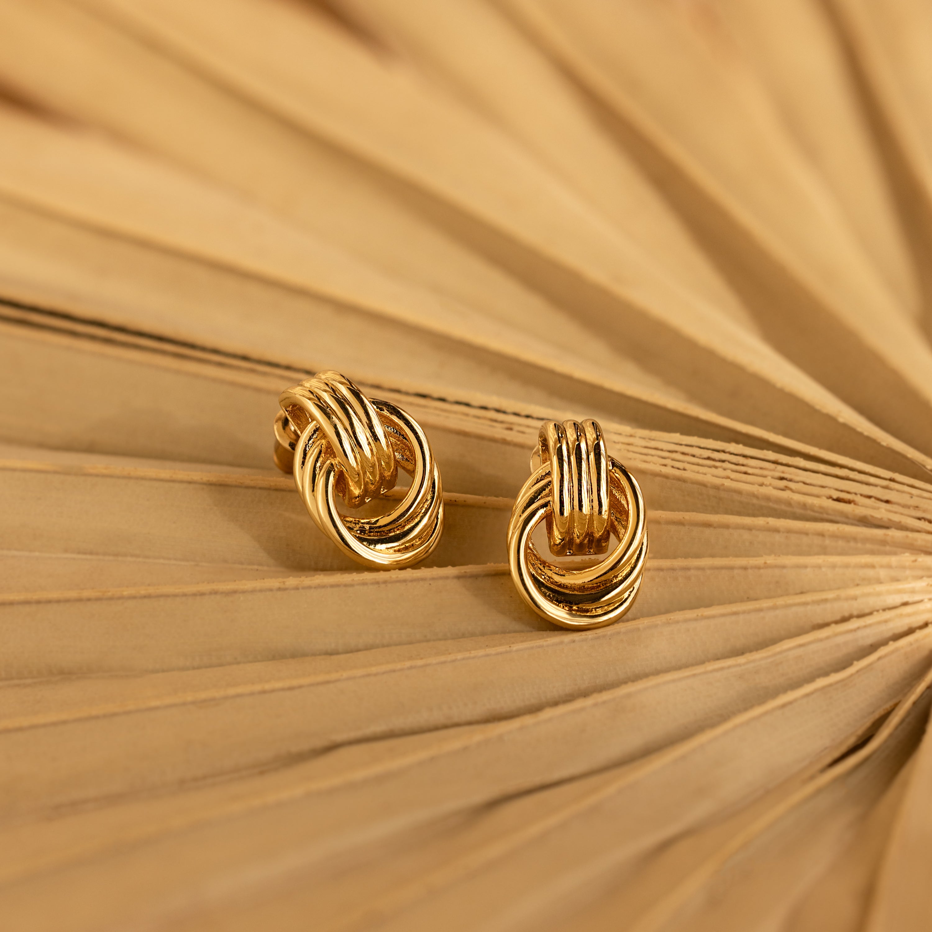 Pair of Mini Triple Knot Earrings styled on the folds of a tan paper fan, showcasing their shiny gold finish.