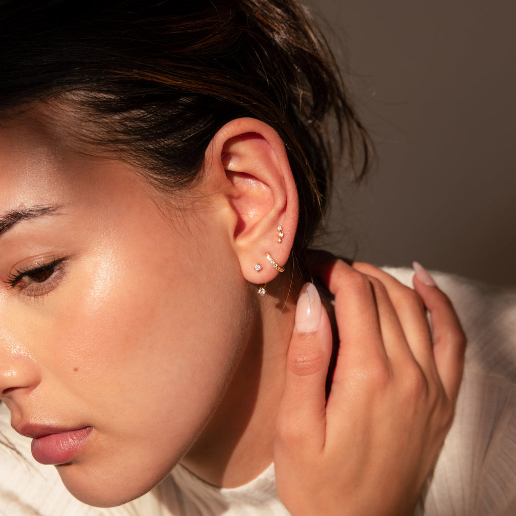 Wearing a white top in warm lighting, a person touches their ear adorned with multiple earrings from the Diamond Earrings Set in 18K Gold, including a sparkling pave huggie hoop.