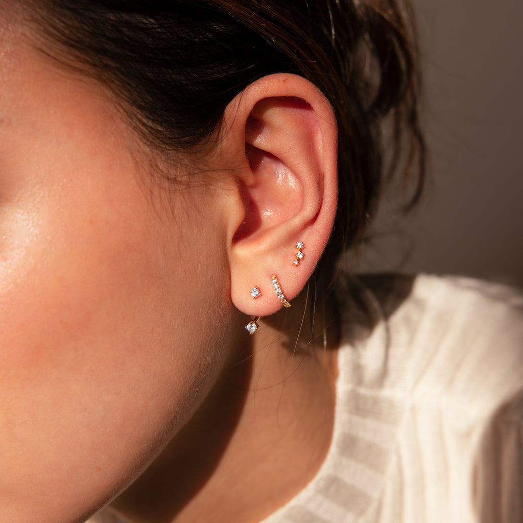 Close-up of a woman's ear wearing the Diamond Earrings Set in 18K Gold, featuring sparkling pavé huggie hoops and delicate studs styled in multiple piercings.