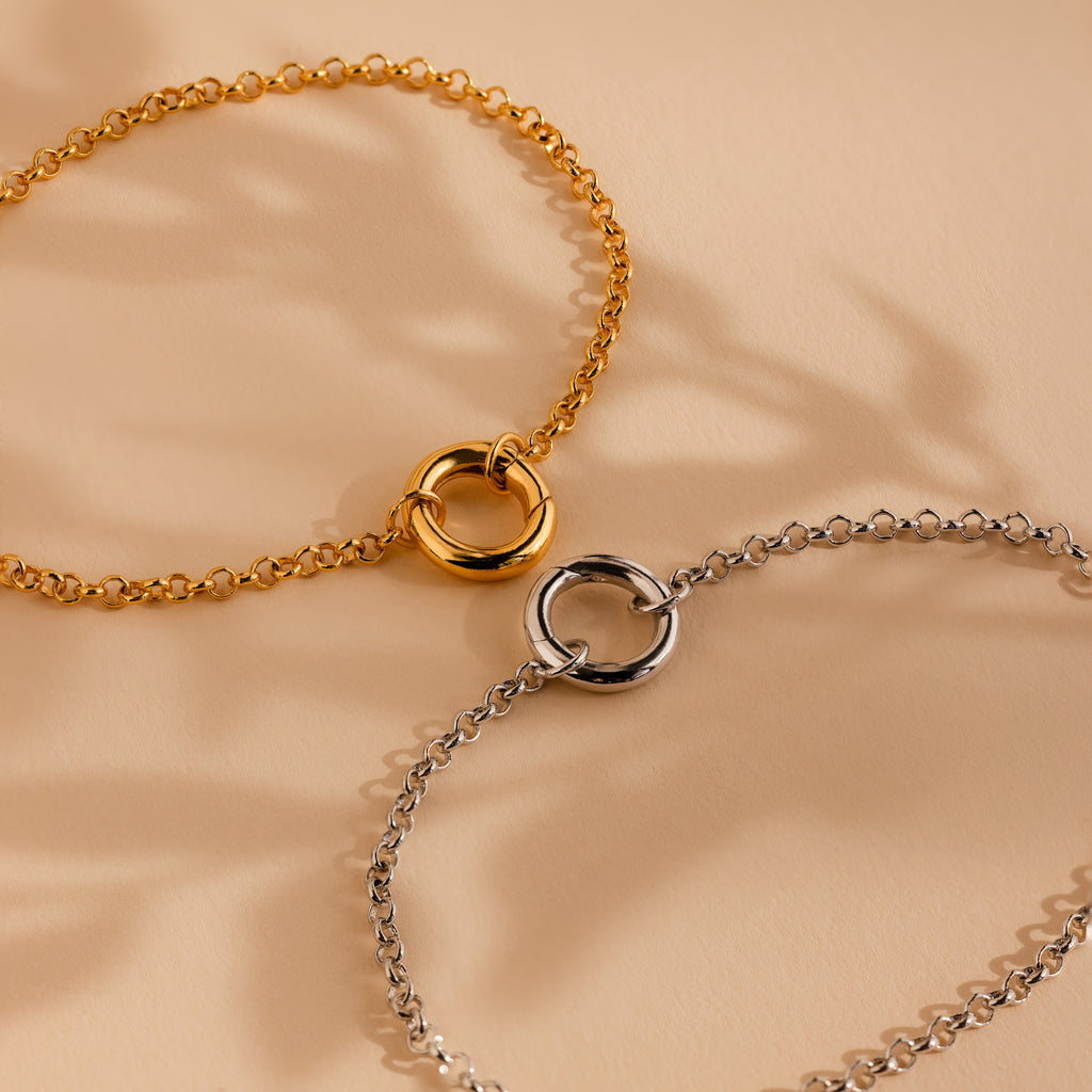 The Rolo Circle Charm Bracelet features a minimalist gold or silver design with a detachable hinged circle clasp, shown on a beige background—ideal for adding charms and personalizing your style.