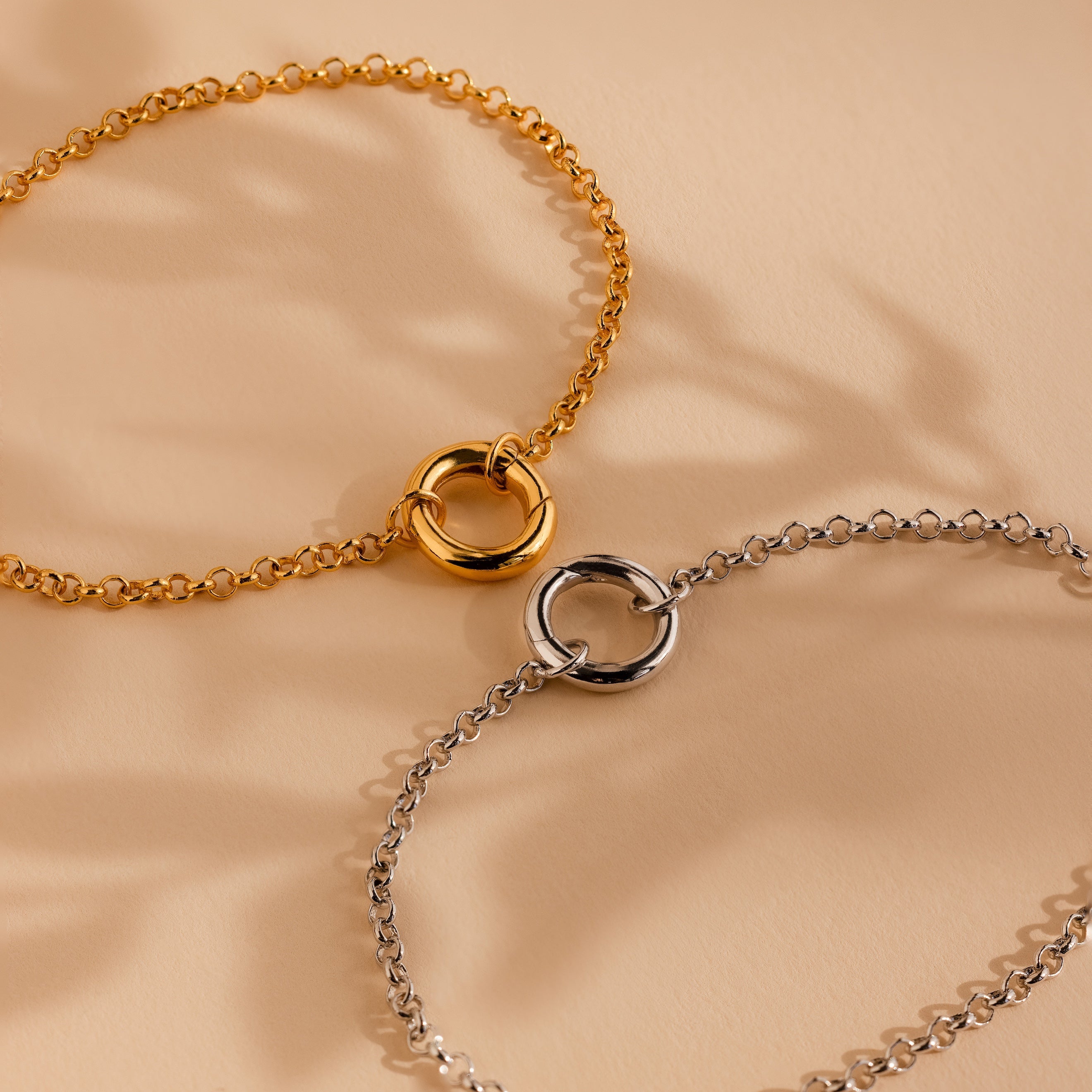The Rolo Circle Charm Bracelet features a minimalist gold or silver design with a detachable hinged circle clasp, shown on a beige background—ideal for adding charms and personalizing your style.