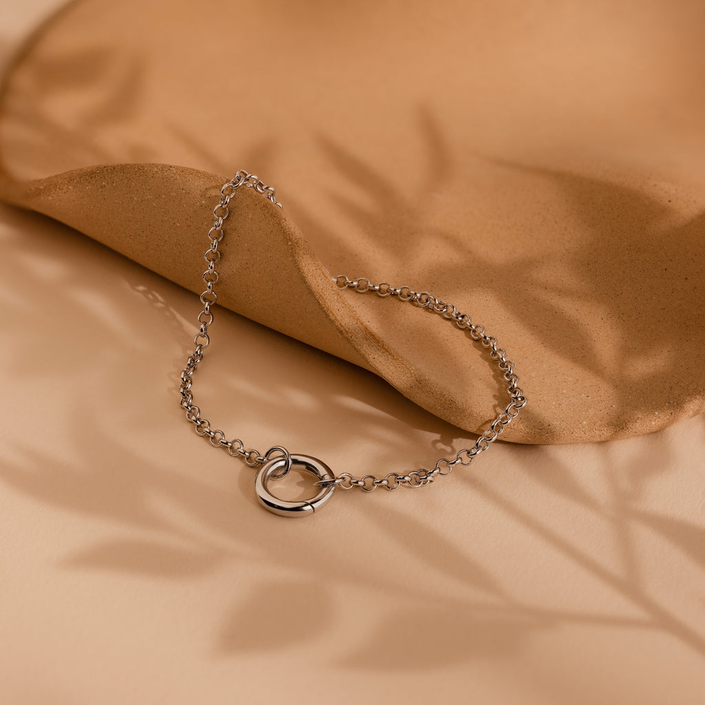 Rolo Circle Charm Bracelet in White Gold with interlocking rings on a beige surface and soft leaf shadows. The detachable hinged circle allows you to easily add charms for your own personalized style.