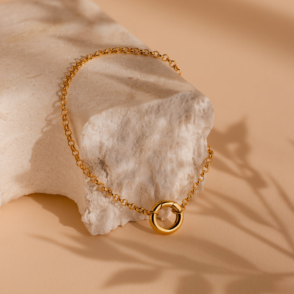 A Rolo Circle Charm Bracelet draped over a textured, light stone on a beige surface.