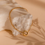 A Rolo Circle Charm Bracelet draped over a textured, light stone on a beige surface.