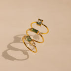 The Agate Emerald Ring in 18K Gold, along with three other gold rings featuring green gemstones, appears to float above a beige background, casting soft shadows.