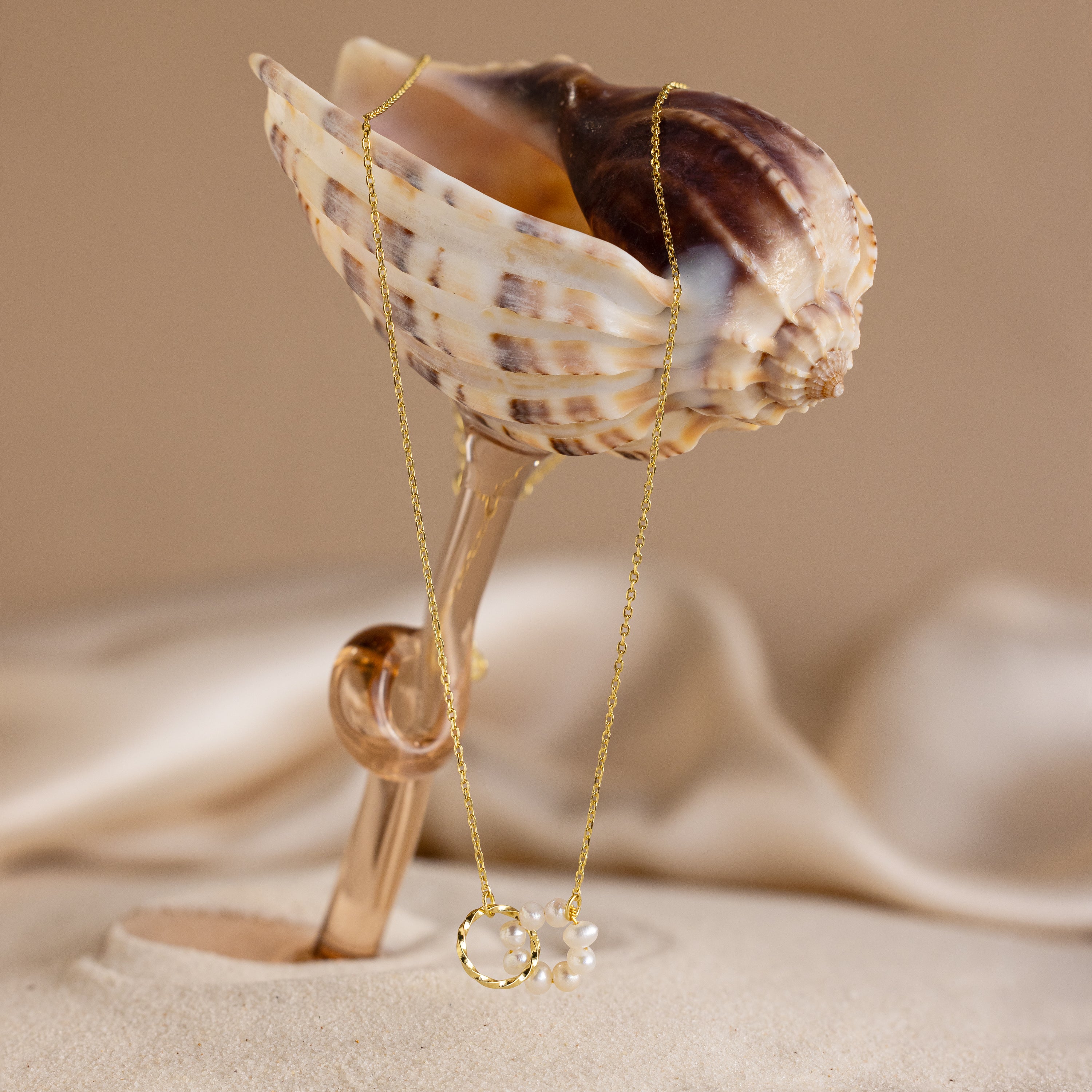 The Interlocking Pearl Necklace in 18K Gold is showcased with pearls draped over a seashell on sandy, neutral fabric.