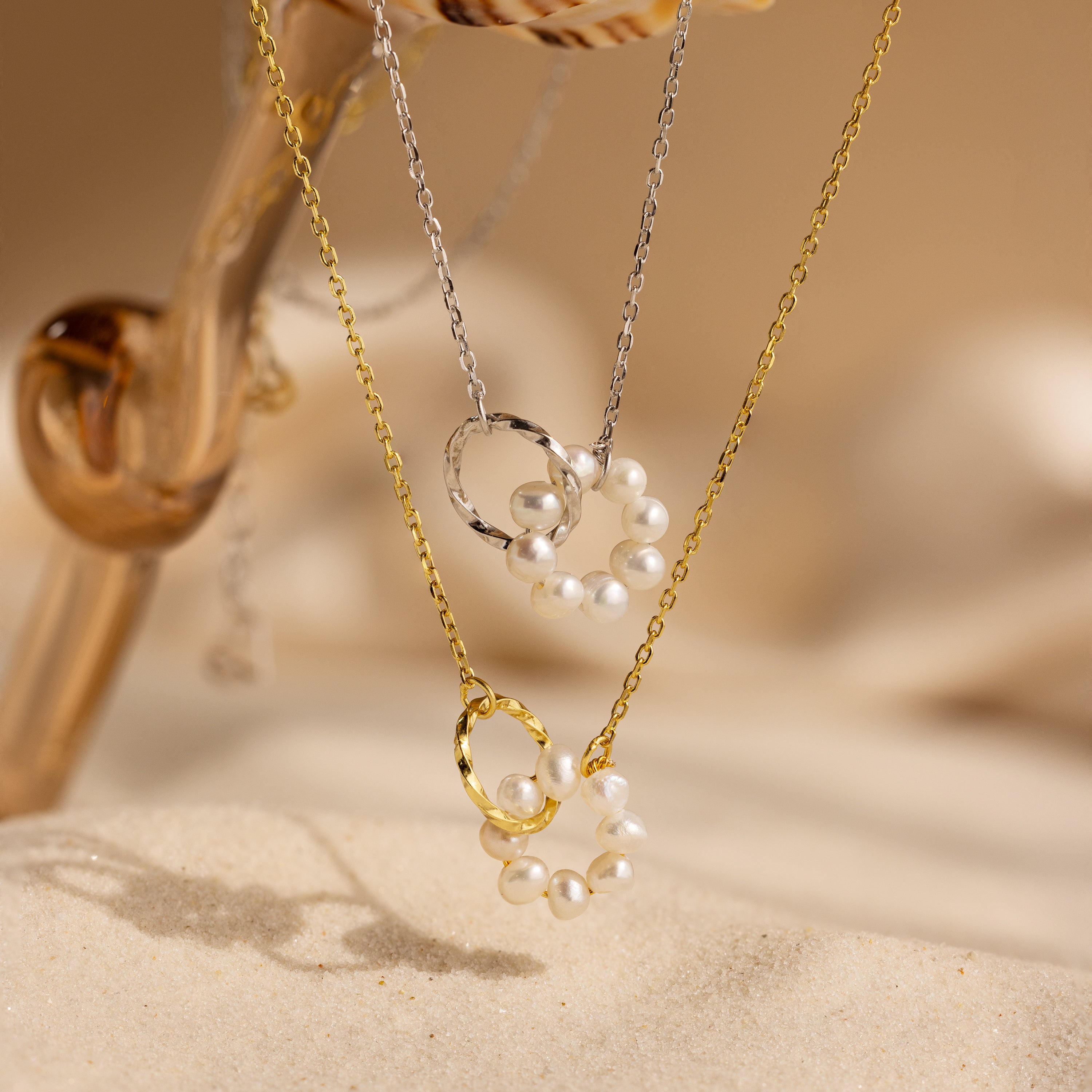 The Interlocking Pearl Necklace, featuring circular pendants and beaded pearl charms, is elegantly displayed on sand and glass.