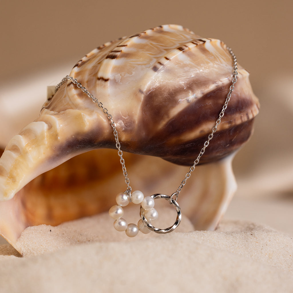 The Interlocking Pearl Necklace is displayed draped over a seashell on sand, set against a beige background.