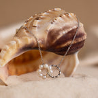 The Interlocking Pearl Necklace is displayed draped over a seashell on sand, set against a beige background.