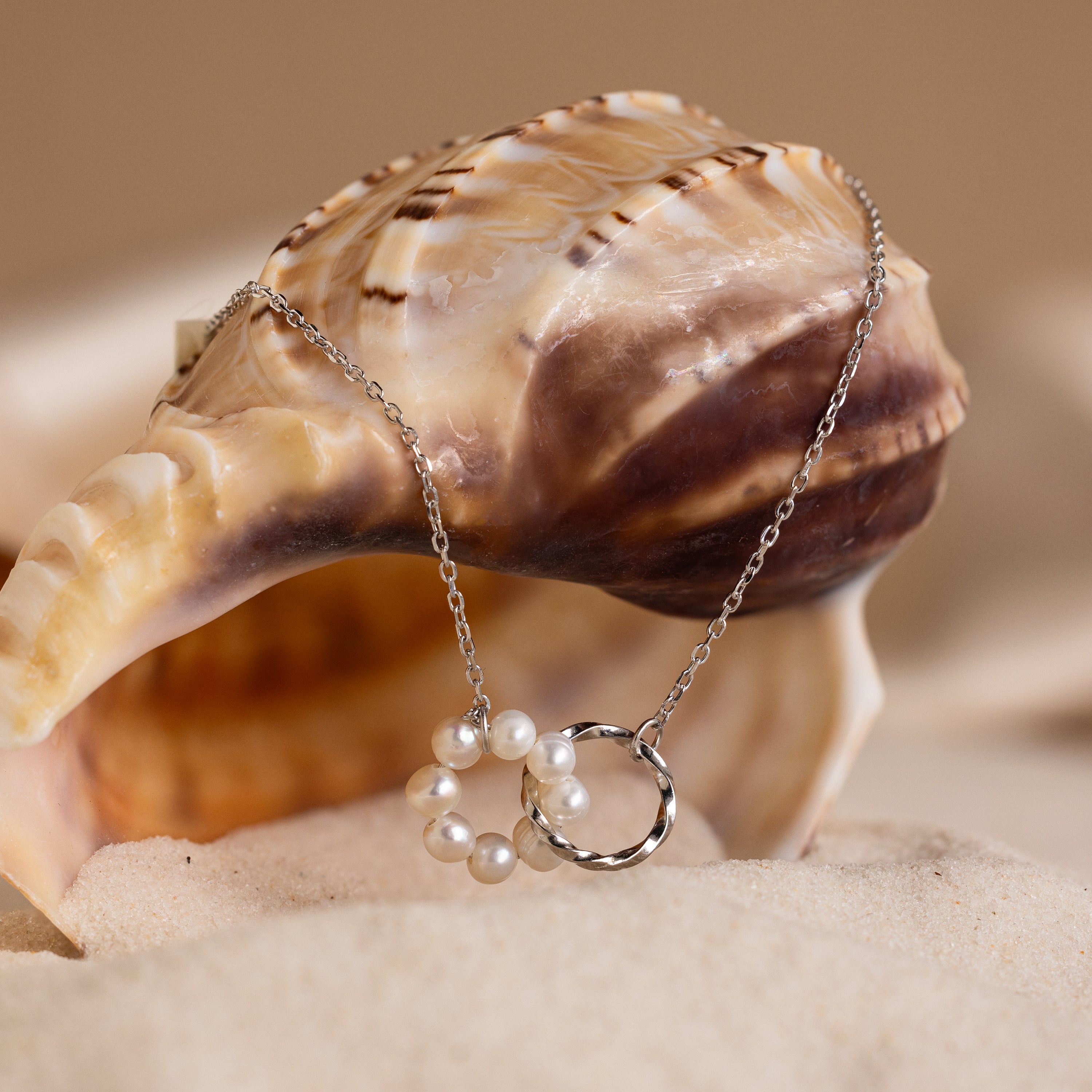 The Interlocking Pearl Necklace is displayed draped over a seashell on sand, set against a beige background.